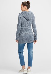 Hoodie lungo blu melange con coulisse, vestibilità slim e vita con cintura, abbinato a jeans tapered azzurri chiari e sneakers bianche.