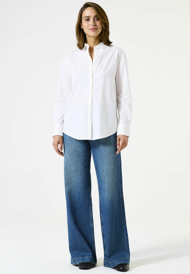 Vrouw staat rechtop, draagt een witte blouse met knopen, wijdvallende blauwe jeans met een subtiel gespetterd patroon en zwarte schoenen, tegen een effen witte achtergrond.