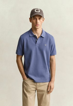 Jeune homme portant un polo bleu, un pantalon beige et une casquette grise avec la lettre G, debout avec une main dans la poche devant un fond uni.