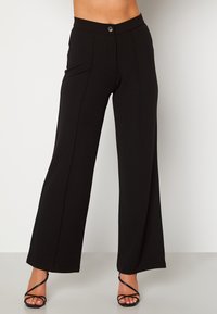 Zwarte wijde broek van gladde stof. Voorzien van een sluiting met een knoop aan de voorkant, plooien en een getailleerde pasvorm. Schoeisel niet inbegrepen.