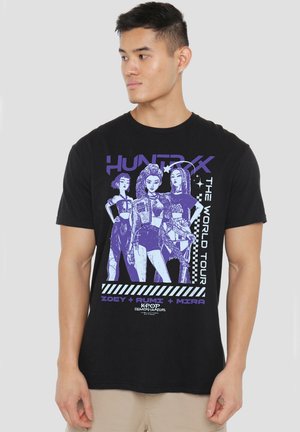 Young man wearing black T-shirt with purple and white graphic of three female characters and text "Hunter X The World Tour" and "Zoey + Rumi + Mira."