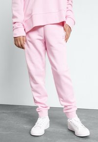 Rosa sweatpants gjorda av mjukt material, med ribbade muddar och en liten logodetalj. I kombination med vita sneakers på ett strukturerat golv.