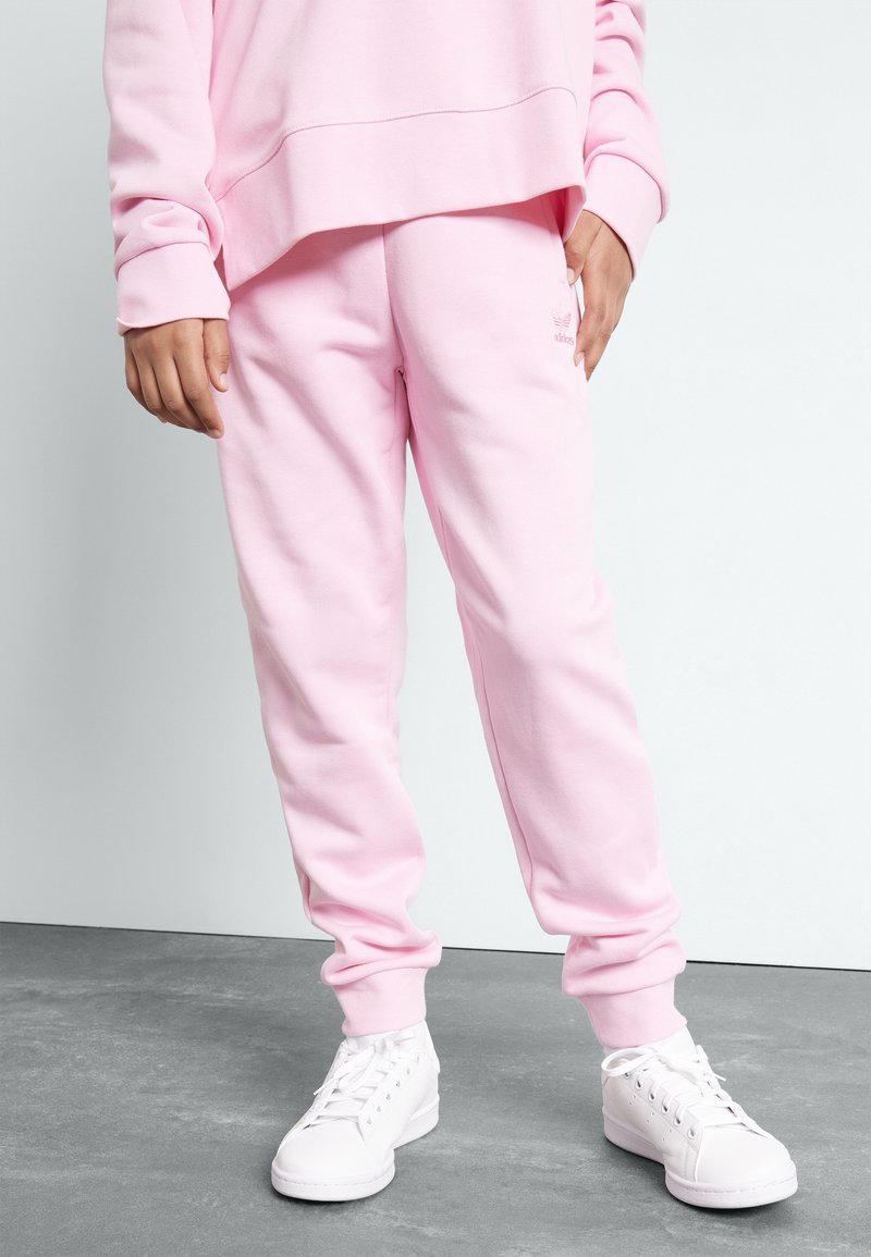 Rosa sweatpants gjorda av mjukt material, med ribbade muddar och en liten logodetalj. I kombination med vita sneakers på ett strukturerat golv.