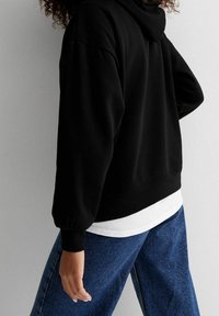 Sudadera negra con capucha y un corte relajado, con puños y dobladillo acanalados. Lleva una camisa blanca debajo y está combinada con jeans de denim azul.