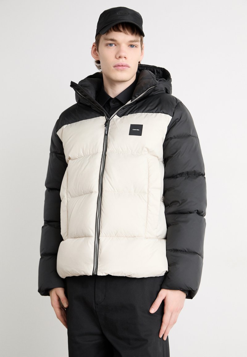 Puffervest med farveblokdesign i sort og creme, med lynlås, hætte og et firkantet mærkelogo på brystet.