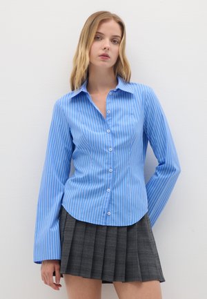 Camicia button-up a righe azzurre con maniche lunghe; presenta un colletto e un orlo curvo. Abbinata a una gonna plissettata grigia.