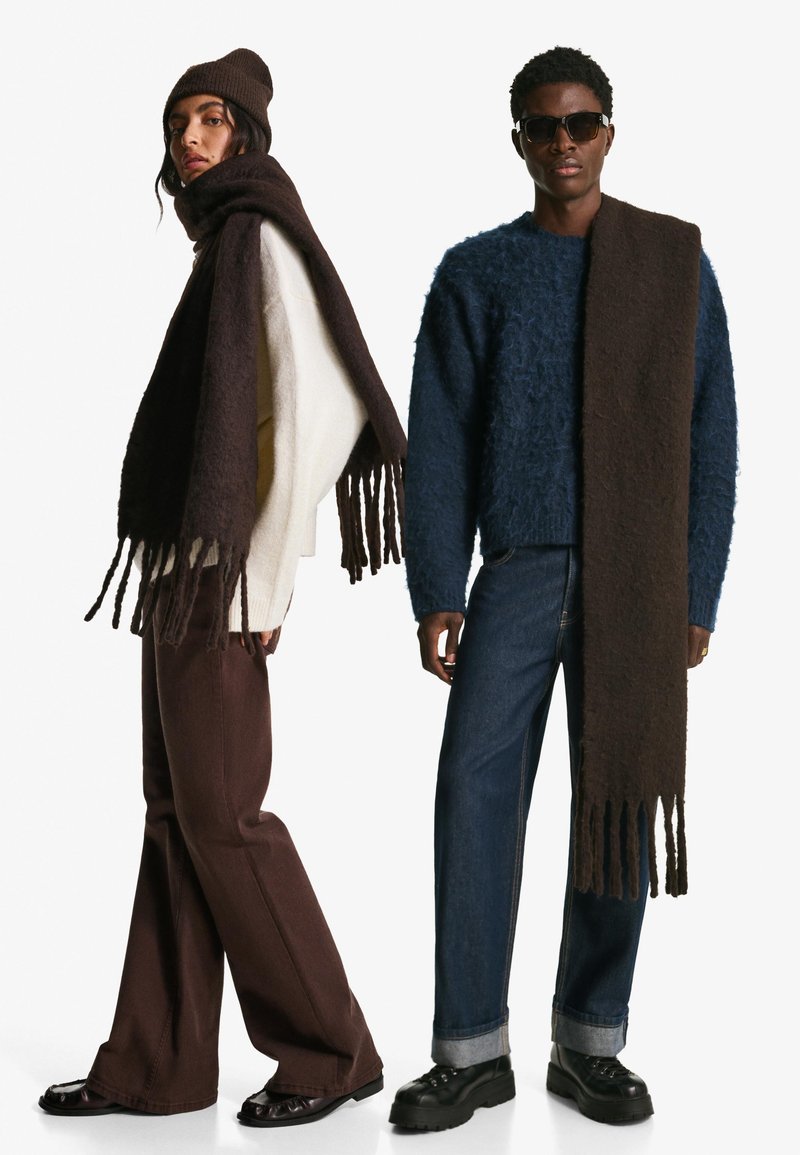 To modeller bærer tøj: en løstsiddende cremefarvet sweater, et mørkebrun tørklæde med frynser, mørkebrun bukser og en fuzzy blå sweater med denim.