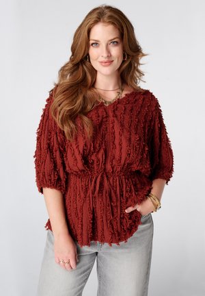 Vrouw met lang rood haar, een gestructureerde roestrode blouse met een tailleband en lichtgrijze spijkerbroek, glimlachend met één hand in haar zak.
