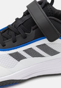 Scarpa sportiva in bianco e nero con tomaia in rete, dettagli blu e cinturino a strappo. Suola testurizzata e design a righe sul lato.