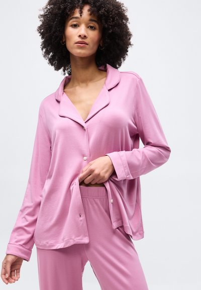 Γυναίκα με σγουρά μαλλιά που φοράει ρόμπα πιτζάμα μακριάς μανσέτας σε ροζ χρώμα, με φαρδιά εφαρμογή και ταιριαστό παντελόνι, πόζα σε απλό φόντο.