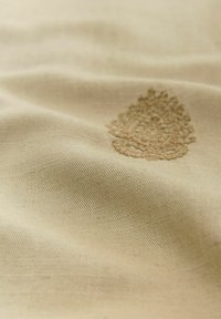 Beiger Baumwollstoff mit strukturiertem Oberfläche und einem floralen Spitzenstickdetail in einem hellbraunen Farbton in der unteren linken Ecke.
