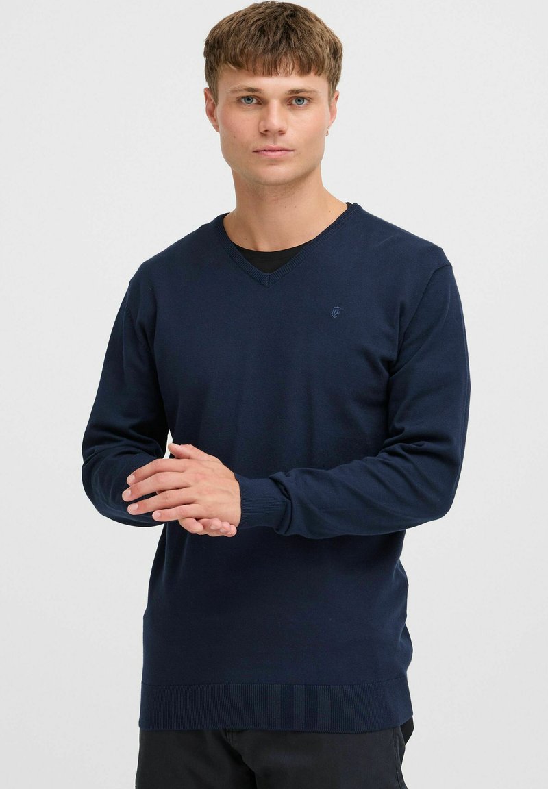 Granatowy sweter w serek wykonany z miękkiej dzianiny, z ribbrowymi mankietami i hemem oraz dyskretnym logo po lewej stronie klatki piersiowej. Prosty, elegancki design.