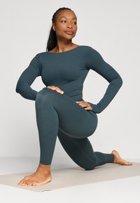 Czarny zielony zestaw odzieży aktywnej z długimi rękawami i legginsami, wykonany z elastycznego materiału. Model przyklęka na macie do jogi, prezentując swoją formę i elastyczność.