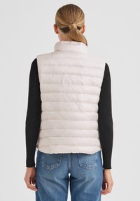 Ärmellose, wattierte Weste in Hellrosa mit Steppmuster, hohem Kragen und glatter Textur, kombiniert mit einem schwarzen Langarmshirt und blauen Jeans.