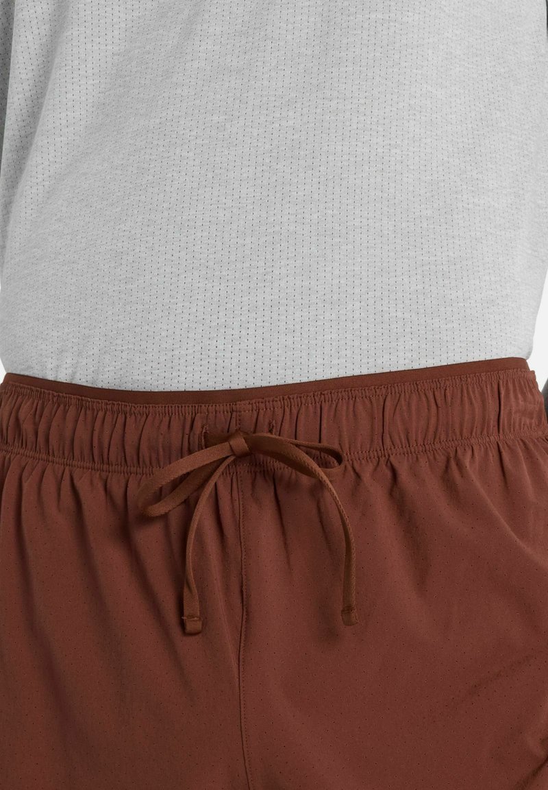 Korte bruine short met een elastische tailleband, voorzien van een trekkoord en een gestructureerd oppervlak, gecombineerd met een lichtgrijs shirt.