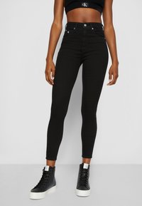 Mujer vestida con jeans skinny de tiro alto negros, top corto negro con el logo de Calvin Klein y zapatillas altas negras con suelas blancas.