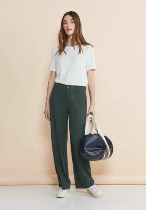 Top bianco a maniche corte, pantaloni larghi verde scuro e borsa circolare blu navy con tracolle bianco sporco. La modella indossa sneakers bianche. Sfondo semplice.