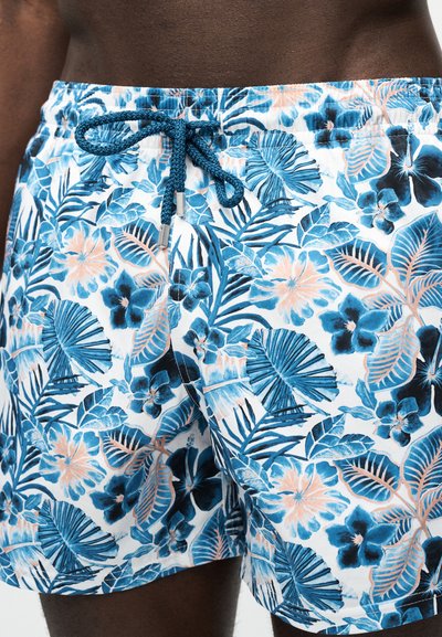 Άτομο με σκούρο δέρμα που φοράει μαγιό με μπλε, λευκό και ροδακινί φλοράλ σχέδιο και μπλε κορδόνι δεμένο σε κόμπο στη μέση.
