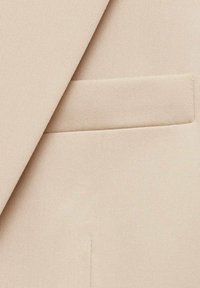 Tissu de costume beige présentant une texture diagonale avec un détail de poche passepoilée simple, offrant une finition lisse et nette.