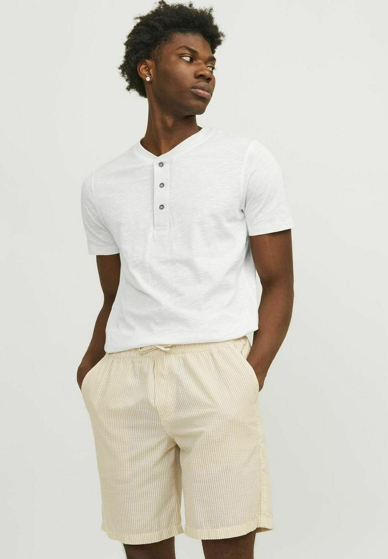 Fehér, rövid ujjú henley ing, három gombbal, világos bézs csíkozott, zsinórral állítható rövidnadrággal viselve, laza szabással és légáteresztő anyagból.