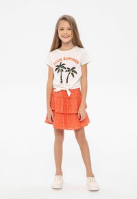 Malá dívka se usmívá, má na sobě bílé tričko "Love Summer" s potiskem palmy, oranžovou sukni s volány a bílé slip-on boty, stojí na bílém pozadí.