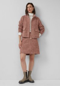 Vrouw in een textuur roze zip-up jack, bijpassende rok, witte coltrui en taupe enkellaarsjes, staand tegen een effen lichte muur.