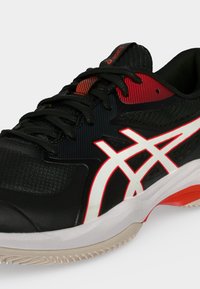 Μαύρο και κόκκινο αθλητικό παπούτσι ASICS με λευκό λογότυπο, μαύρα κορδόνια και λευκή και πορτοκαλί σόλα σε απλό φόντο.