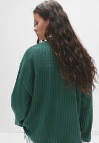 Dlhý zelený rebrovaný sveter s voľným strihom a textúrovaným povrchom, so zvislými pruhmi a voľne priliehajúcimi rukávmi.
