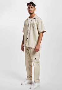Beige short-sleeve button-up shirt with a chest pocket, paired with matching cargo pants featuring side pockets and black buttons. White sneakers.