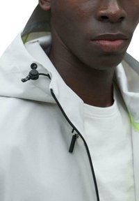 Chaqueta impermeable gris claro con cremallera negra y capucha ajustable. Presenta un sutil acento neón en el interior del cuello y una textura suave.