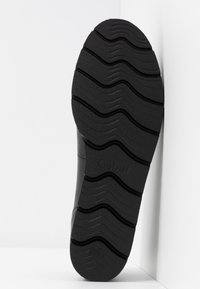 Semelle de chaussure noire avec motif de bande de roulement ondulée et rainurée pour l'adhérence, positionnée verticalement sur un fond d'angle blanc.