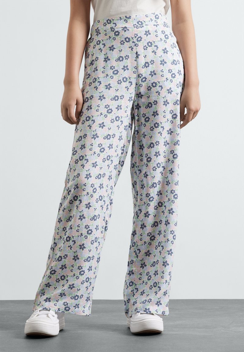Pantalons larges floraux en tissu léger, avec un motif pastel de fleurs bleues et vertes, assortis à des baskets blanches et un haut sobre.