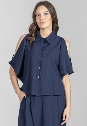 Kobieta o falowanych brązowych włosach, ubrana w granatową bluzkę z odkrytymi ramionami zapinaną na guziki oraz pasującą spódnicę, stojąca z jedną ręką w kieszeni.