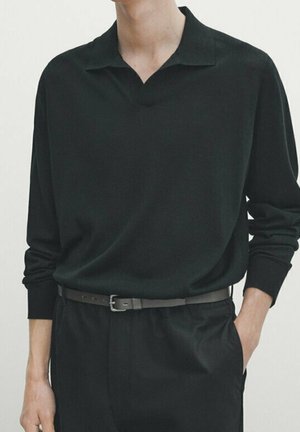 Personne portant une chemise noire à manches longues avec un col souple rentrée dans un pantalon noir avec une fine ceinture noire.