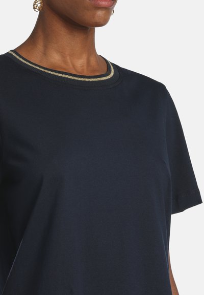 Sötétkék rövid ujjú felsőt viselő személy arany csíkos, kerek nyakkivágással és arany filigrán fülbevalóval, egyszerű háttér előtt.