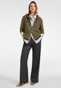 Olivengroene getextureerde hoodie met een ritssluiting aan de voorkant, gecombineerd met een gestreept shirt en donkere brede broek. Opvallende details zijn een zwarte rand en zakken.