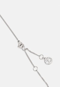 Catena in metallo argento con chiusura a moschettone, caratterizzata da anelli intrecciati e un charm con logo. Texture liscia con finitura lucida.