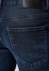 Ciemnoniebieskie dżinsy z tylną kieszenią z detalami w szwie oraz czarną skórzaną naszywką marki w talii. Delikatna struktura na całej powierzchni.