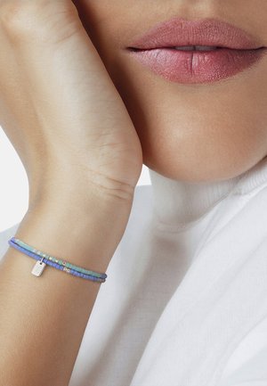 Plano detalle de la parte inferior del rostro y la mano de una persona apoyada en la mejilla, que lleva una camiseta blanca y pulseras de cuentas azules y turquesas con un pequeño encanto de plata.