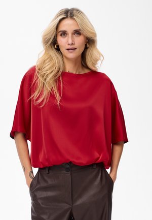 Blonde vrouw met losse golven in haar haar draagt een rode blouse met korte mouwen en donkere leren broeken, met handen in haar zakken, tegen een effen witte achtergrond.