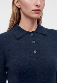 Femme portant un pull duveteux bleu foncé avec un col et trois boutons, montrée des épaules au menton.