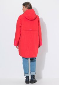 Długa czerwona kurtka przeciwdeszczowa z kapturem, o prostym kroju, z kontrastowymi różowymi akcentami i gładką, wodoodporną fakturą.