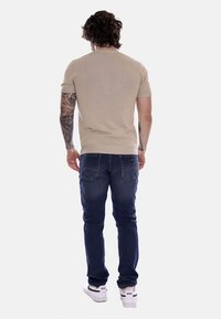 Camicia beige a maniche corte, design aderente, abbinata a jeans blu scuro. Le scarpe sono bianche con marchio Nike nero. Vista posteriore mostrata.