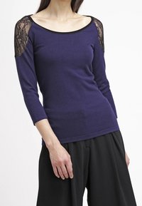 Marineblaues Oberteil mit Drei-Viertel-Ärmeln und schwarzen Spitzenakzenten an den Schultern, rundem Ausschnitt und weicher Textur. Kombiniert mit lockeren schwarzen Hosen.