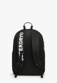 Zaino nero con spallacci imbottiti, dotato di una maniglia superiore e di un grande logo bianco sul davanti e sugli spallacci. Tessuto durevole.