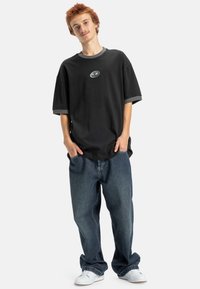 T-shirt noir surdimensionné avec des bordures grises, arborant un petit logo sur la poitrine. Porté avec un jean en denim bleu ample et des baskets blanches.