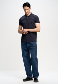 Tamsiai pilka medvilninė polo marškinėliai trumpomis rankovėmis, derinami su mėlynomis džinsinėmis kelnėmis. Vyras stovi neutraliame fone, rankos sukryžiuotos. Juodi batai.