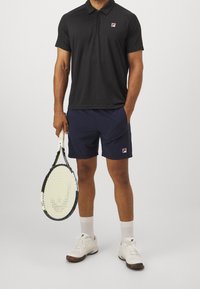 Czarna koszulka polo z kołnierzykiem na zamek, granatowe szorty, białe buty tenisowe oraz rakieta tenisowa z wzorzystym uchwytem. Logo Fila na koszulce i szortach.
