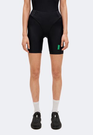 Personne portant un cuissard de cyclisme noir taille haute, des chaussures de sport noires et des chaussettes blanches montantes, debout devant un fond clair uni.