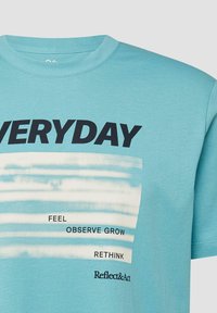 T-shirt bleu clair avec texte noir "EVERYDAY" et les mots plus petits "FEEL", "OBSERVE GROW", "RETHINK" et "Reflect&Act" sur des rayures horizontales blanches.